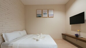 Ein ordentlich bezogenes Bett mit weißer Tagesdecke und Kissen, ein Flachbildfernseher an der Wand sowie drei gerahmte Bilder oberhalb des Bettes.