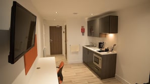 A compact kitchen with modern appliances, a dining area with a white table and orange chairs, and a TV mounted on the wall.