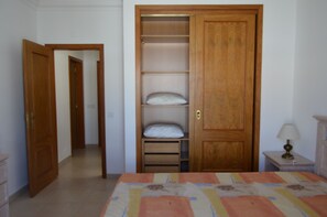 A bedroom with a bed, a wooden wardrobe, a bedside table with a lamp, and a door leading to another room.