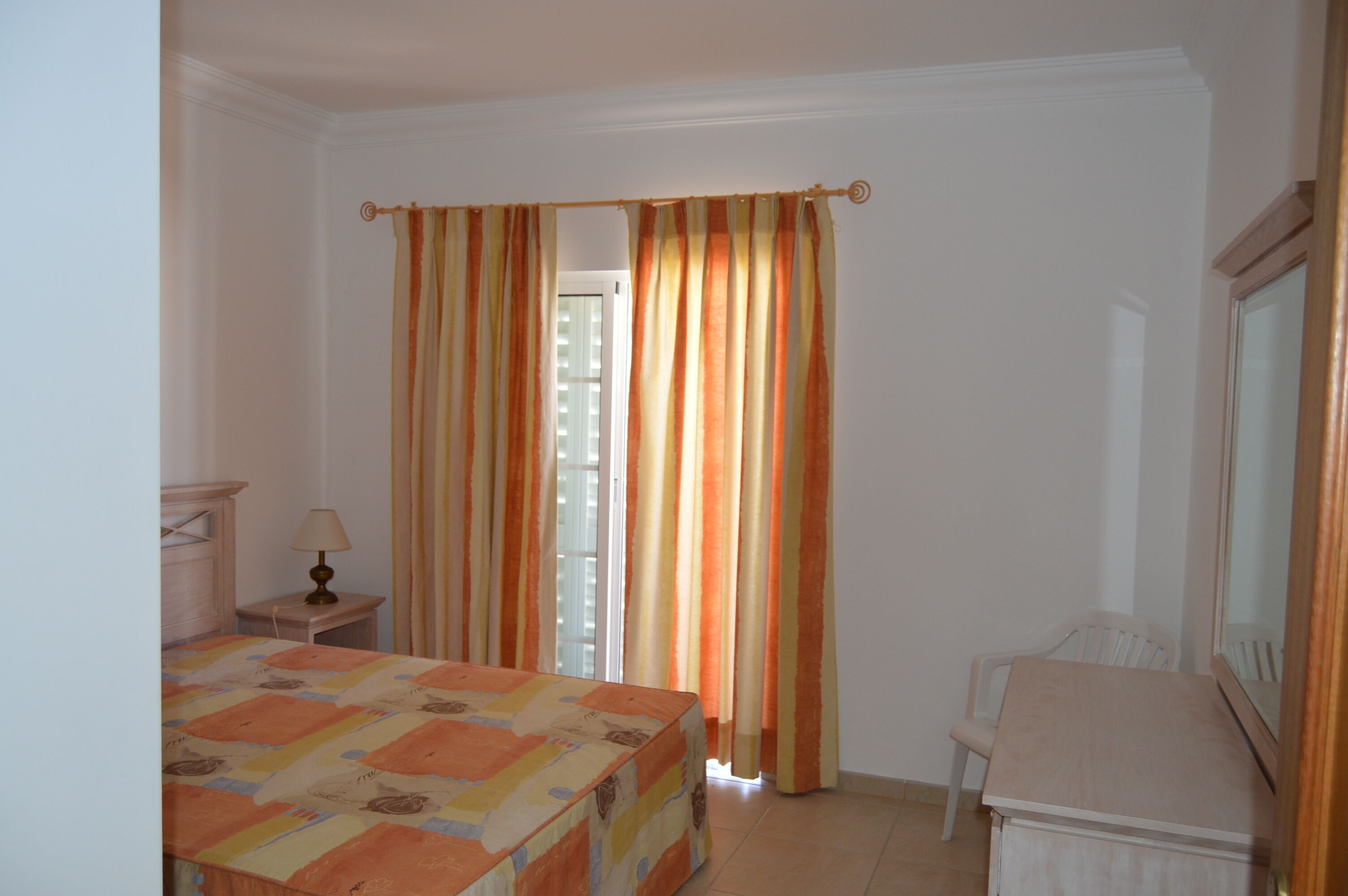 Un dormitorio con cama, mesita de noche, silla y cortinas.