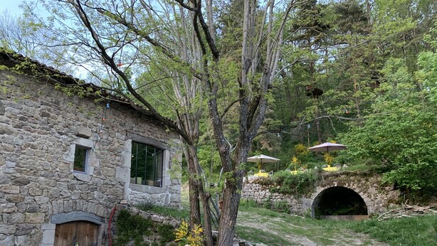 Un bâtiment rustique en pierre, doté d’une petite fenêtre, d’une porte et d’une terrasse en gravier avec des sièges d’extérieur.