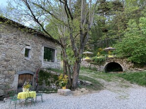 Un bâtiment rustique en pierre, doté d’une petite fenêtre, d’une porte et d’une terrasse en gravier avec des sièges d’extérieur.