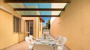 Un área de comedor al aire libre techada, con una mesa dispuesta para cuatro personas, rodeada de sillas blancas, bajo una cubierta de vidrio.