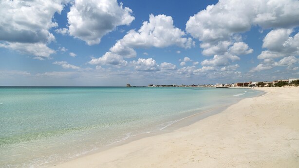 Una playa con agua turquesa cristalina, una orilla arenosa y un pueblo lejano.