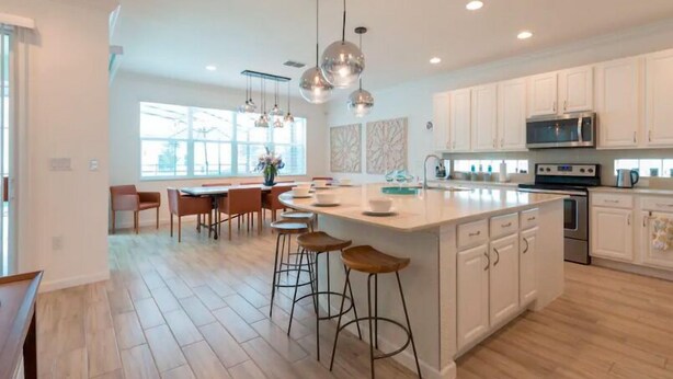 A modern kitchen with white cabinets, a large island with stools, pendant lighting, and a view of the outdoors through large windows. - Brand New Home at Sonoma Resort at Tapestry (Kissimmee)