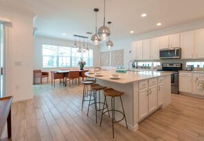 A modern kitchen with white cabinets, a large island with stools, pendant lighting, and a view of the outdoors through large windows. - Brand New Home at Sonoma Resort at Tapestry (Kissimmee)