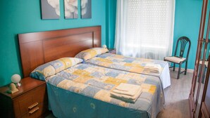 Un dormitorio con dos camas, cabecero de madera, una mesita de noche, una silla y una ventana con cortina translúcida.