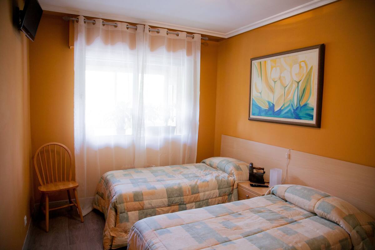 A room with two beds, a wooden chair, a painting on the wall, and a window with curtains.