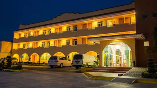 Un hotel con una estructura de varios pisos, una entrada central con marquesina de cristal y un estacionamiento con varios vehículos.