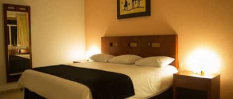 Habitación de hotel con una cama grande, una mesita de noche con lámpara, un cuadro en la pared y un espejo.