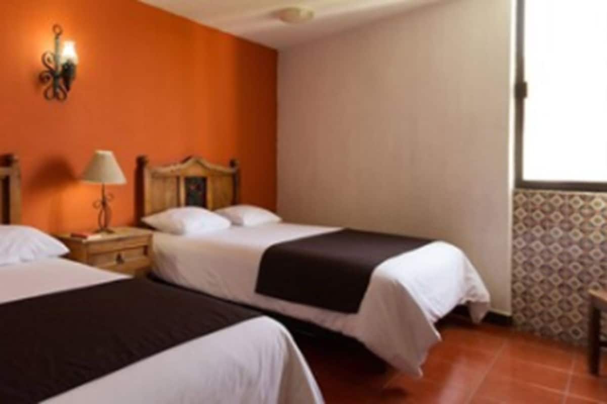 Habitación de hotel con dos camas, una mesita de noche, una lámpara fijada a la pared y una ventana con cortinas estampadas.