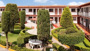 Un patio con setos cuidadosamente recortados, una mesa redonda con sillas, y un edificio de techo con tejas y varias ventanas.