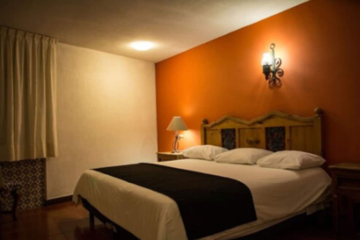 Una habitación de hotel con cama, mesita de noche, lámpara y luz empotrada en la pared.