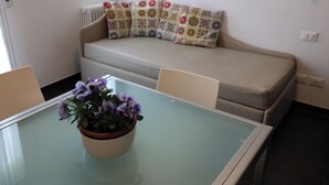 Un petit salon avec une table à plateau en verre, un canapé agrémenté de coussins à motifs et une plante en pot aux fleurs violettes.
