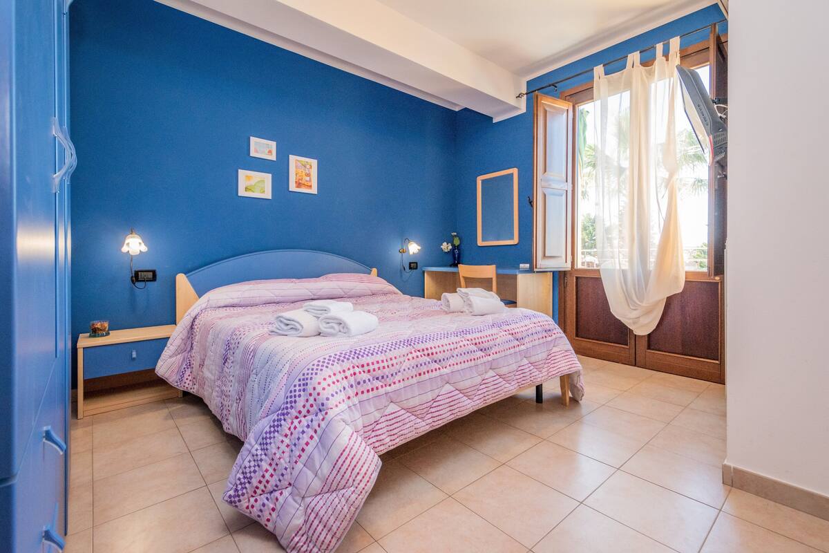 Une chambre à coucher avec un lit, des lampes de chevet, une table de nuit, une fenêtre avec des rideaux et une vue sur des palmiers.