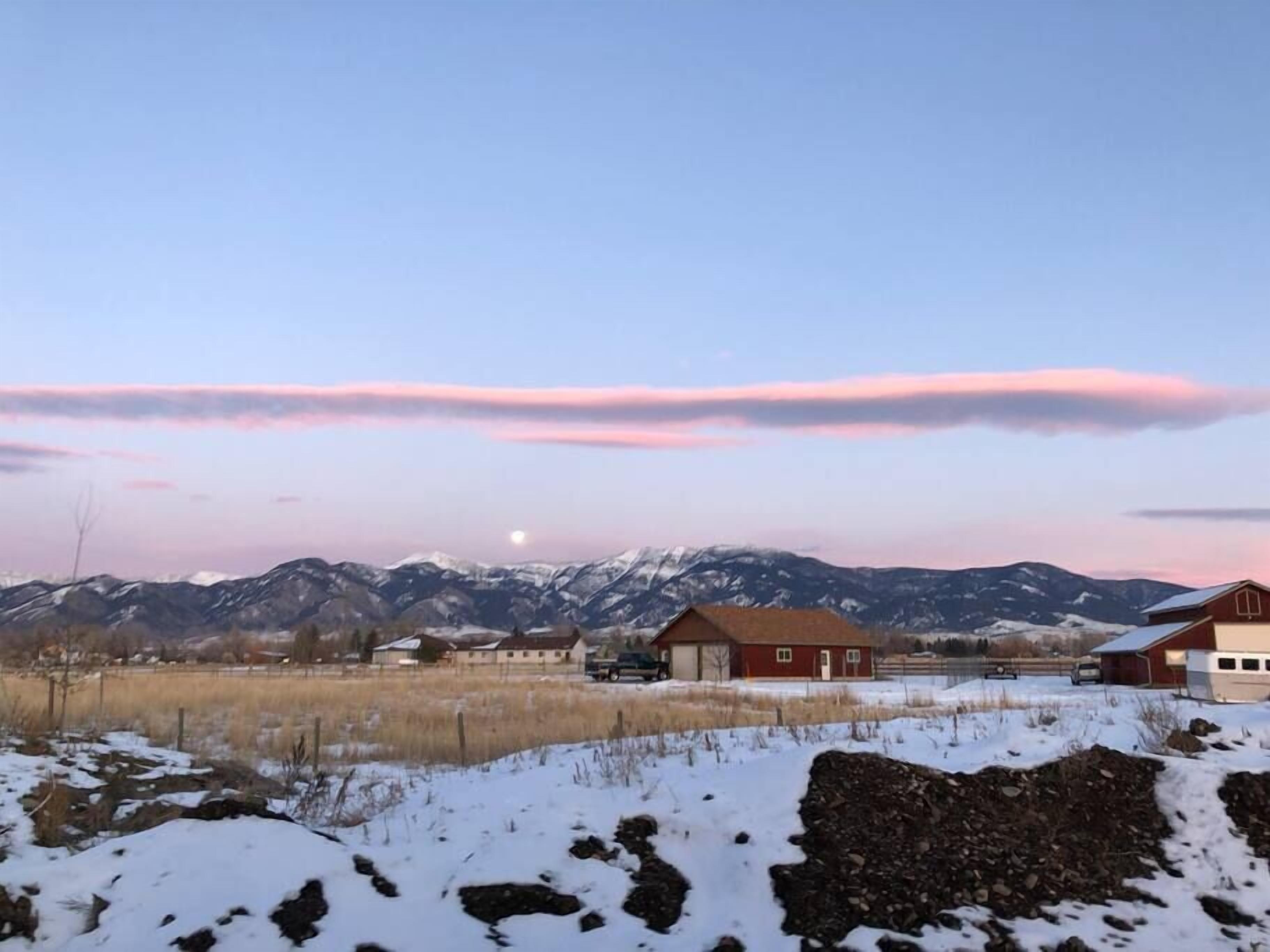 Ett lantligt landskap med en klar himmel, snötäckt mark och en rad röda byggnader, med ett bergskedja i bakgrunden.