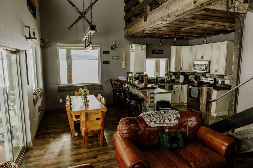 Un salon spacieux avec un canapé en cuir, une table à manger en bois et un coin cuisine doté de meubles blancs et d’un plafond en bois.