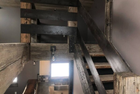 A rustic interior with exposed wooden beams, a staircase, and a kitchen area with a window providing natural light.