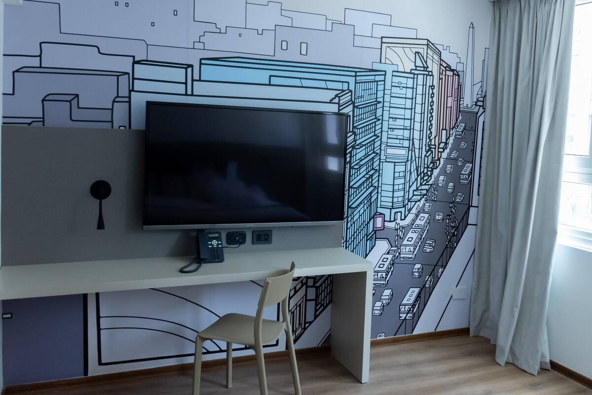 A modern room with a large flat-screen TV on a desk, a chair, and a cityscape mural on the wall.