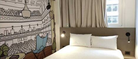 Una habitación de hotel con cama, cabecera, mesita de noche, lámpara, silla, una mesa con una planta y un mural en la pared.