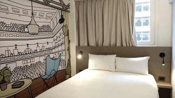 A hotel room with a bed, a headboard, a nightstand, a lamp, a chair, a table with a plant, and a mural on the wall. - ibis Styles Buenos Aires Florida (Buenos Aires)