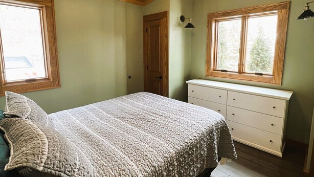 Une chambre avec un plafond en bois, un lit recouvert d’une couverture à motifs, une commode et deux fenêtres.