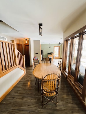 Un comedor amplio con una mesa y sillas de madera, una escalera con barandilla de madera, y ventanas grandes a un lado.