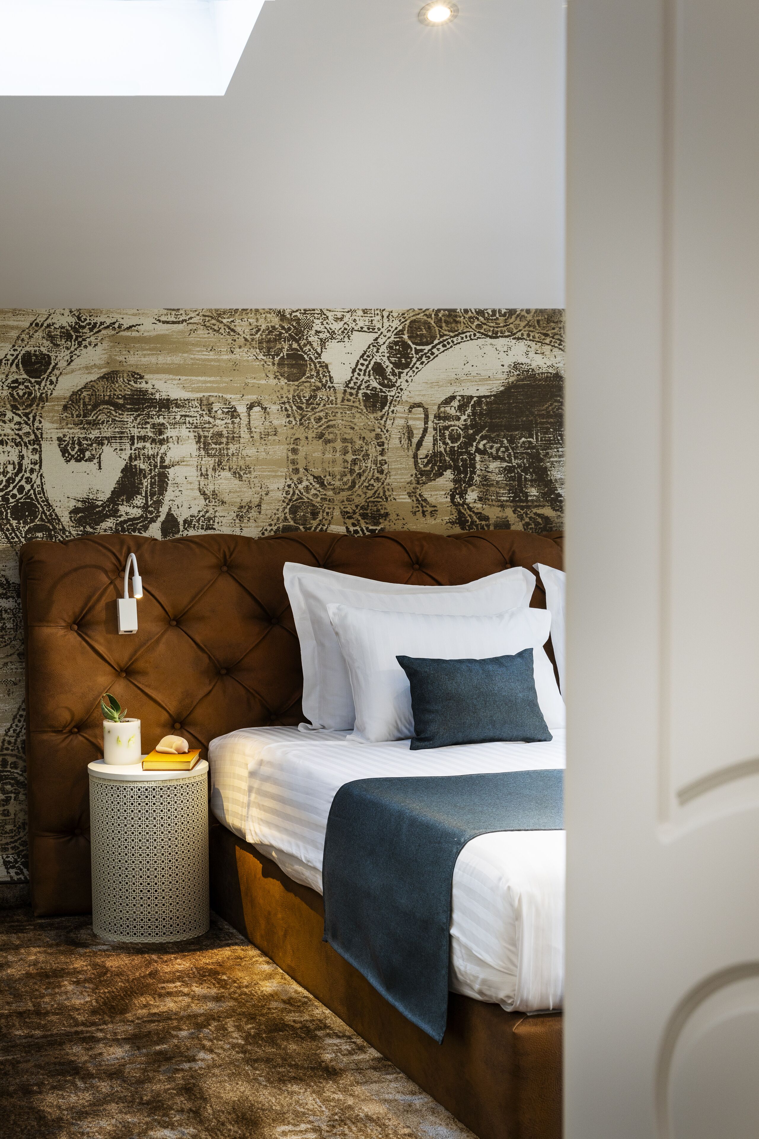 A bedroom with a tufted headboard, a round waste basket, a bedside table with a lamp, a bed with white bedding and a blue throw, and a patterned wallpaper.