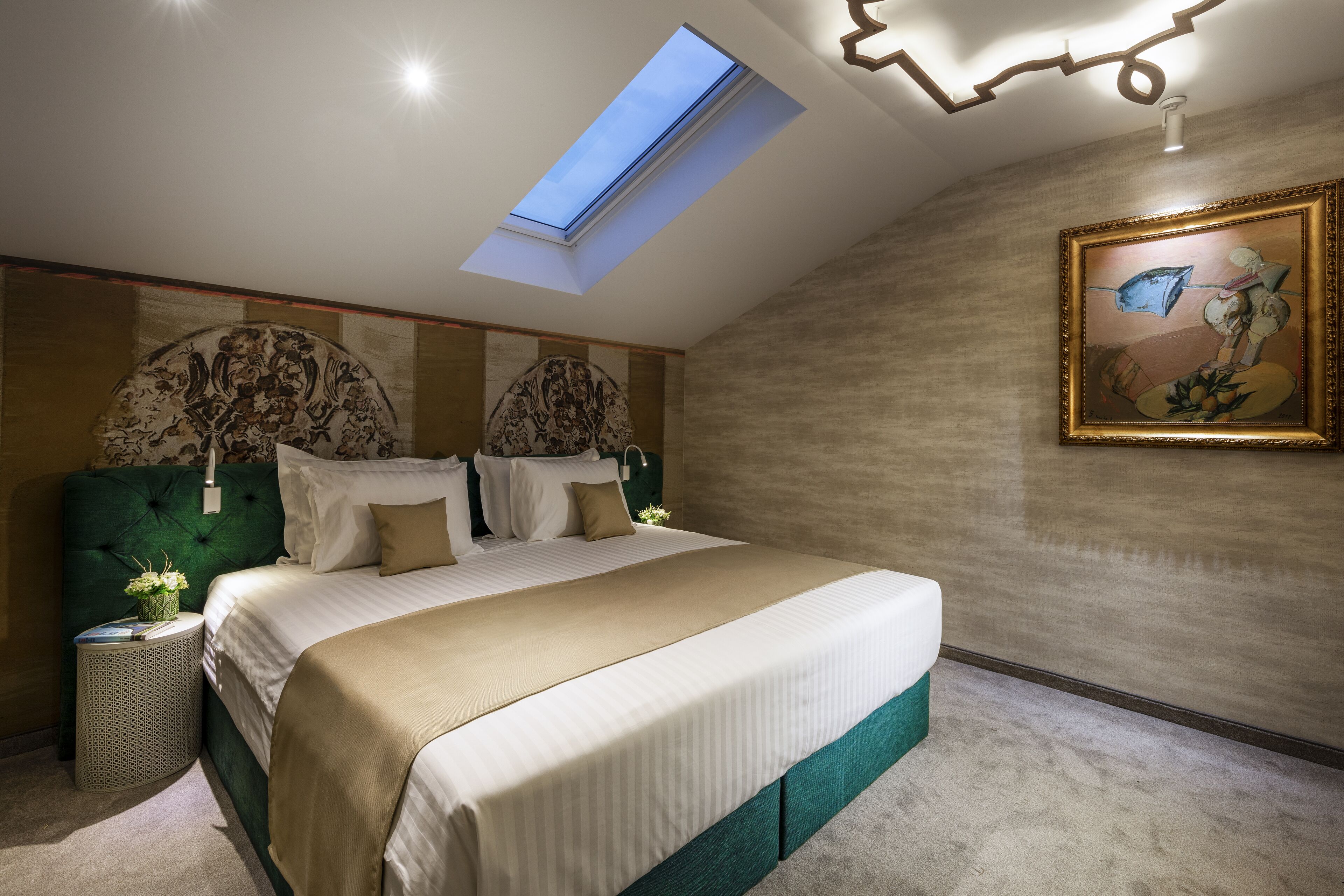 A bedroom with a skylight, a large bed, a painting, and a nightstand with a lamp.