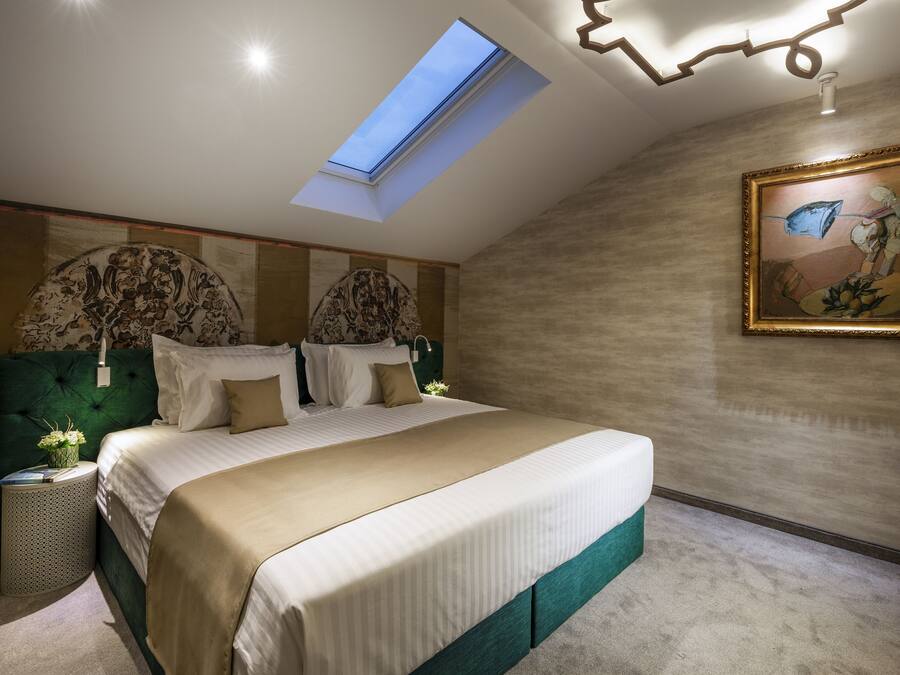 A bedroom with a skylight, a large bed, a painting, and a nightstand with a lamp.