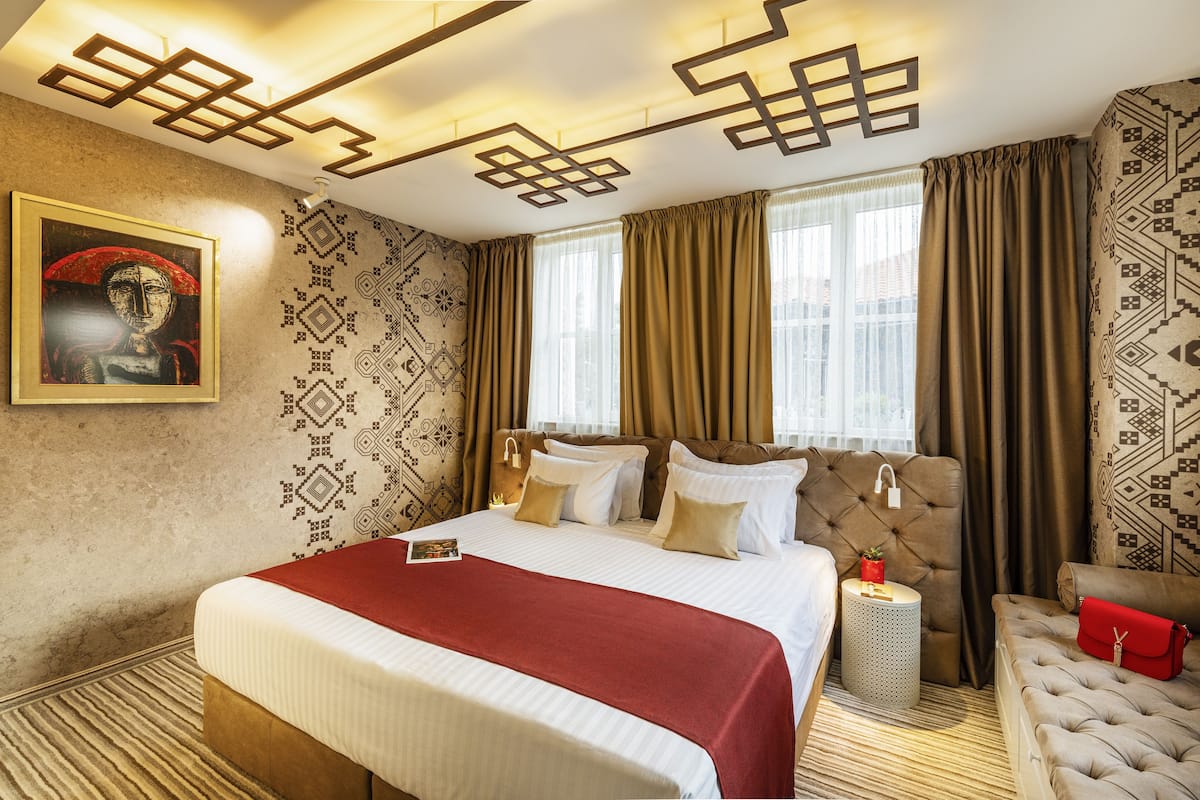 Quarto de hotel com uma cama grande, cabeceira, mesa de cabeceira e uma bolsa vermelha.