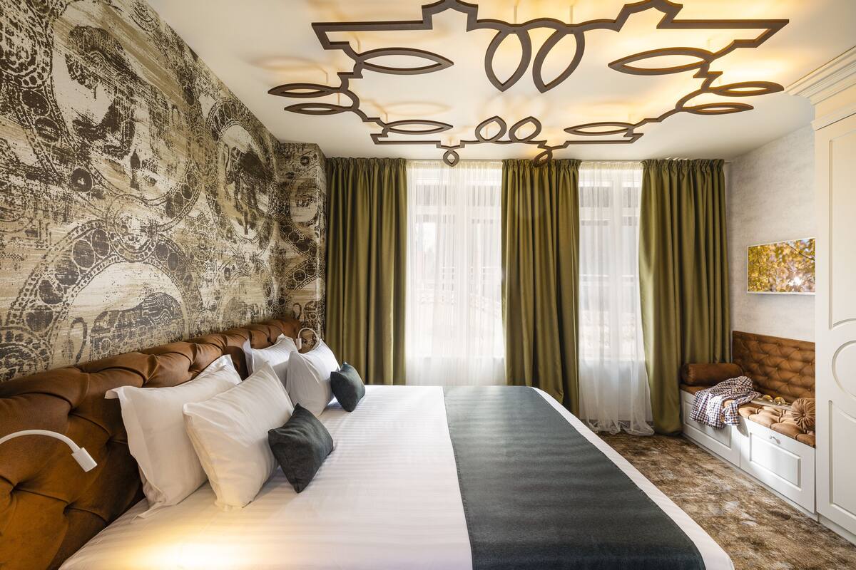 Quarto de hotel com cama grande, cortinas verdes, parede estampada e design de teto singular.