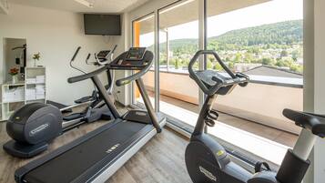 Ein Fitnessraum mit Laufbändern und Crosstrainern, einem an der Wand montierten Fernseher und Blick auf eine Wohngegend durch große Fenster.