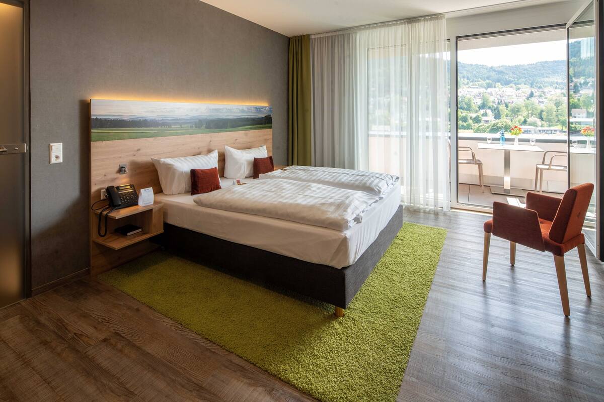 Habitación de hotel con una cama grande, un escritorio con teléfono y vistas a un balcón con sillas.