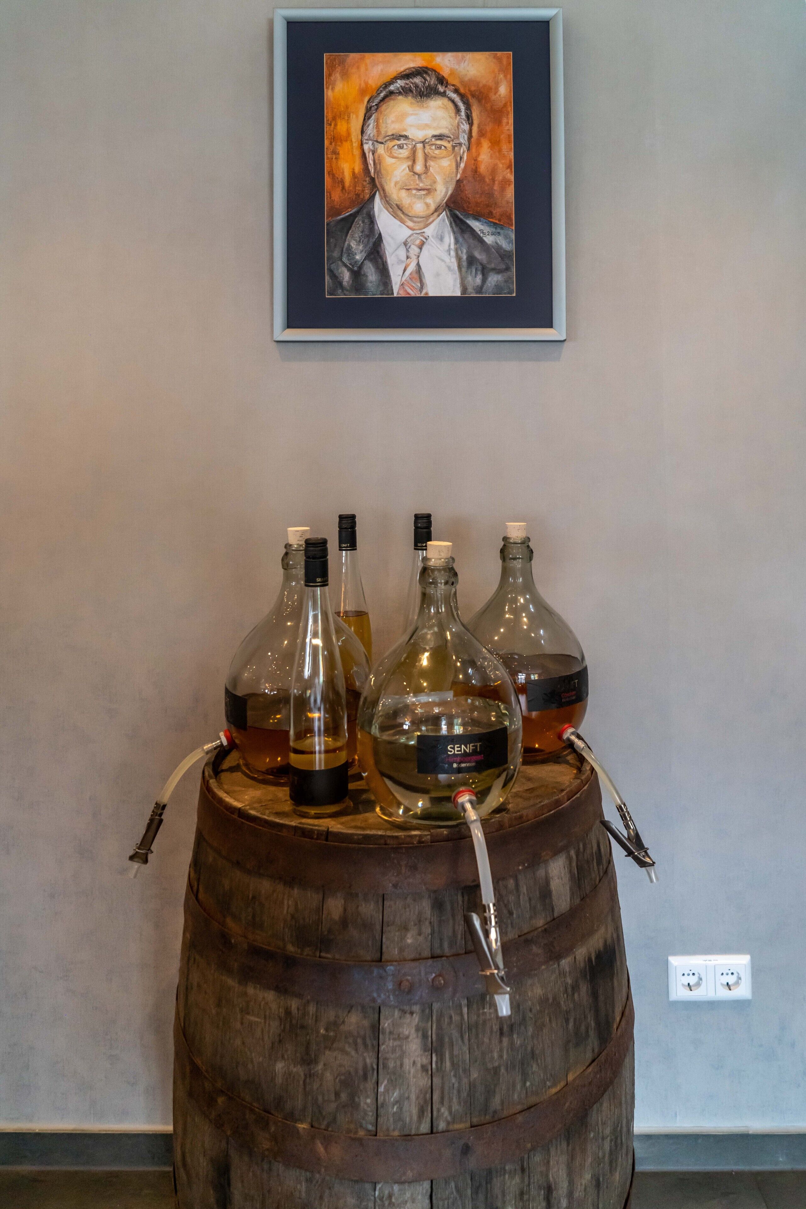 Un barril de madera con botellas de vino y un grifo, un retrato enmarcado en la pared, y un enchufe en la pared.