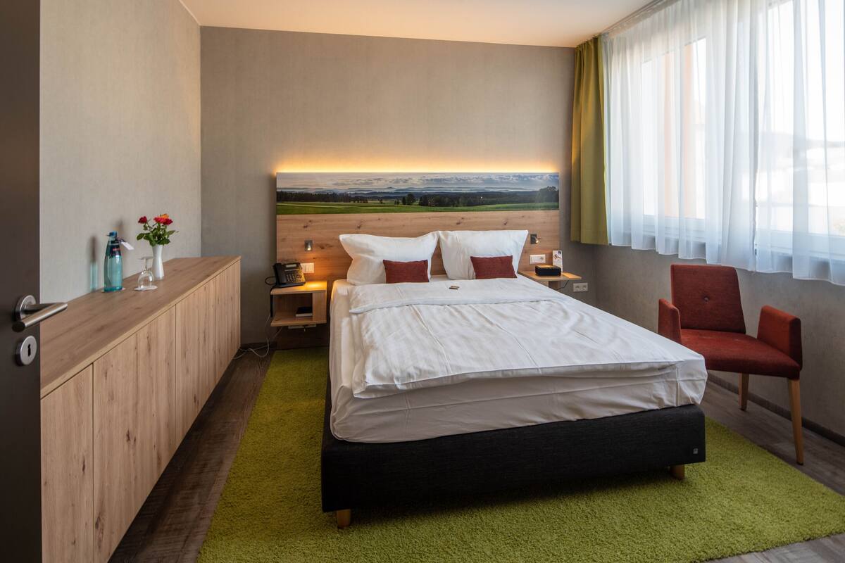 Habitación de hotel con cama, escritorio, silla, mesita de noche, lámpara y ventana con cortinas.