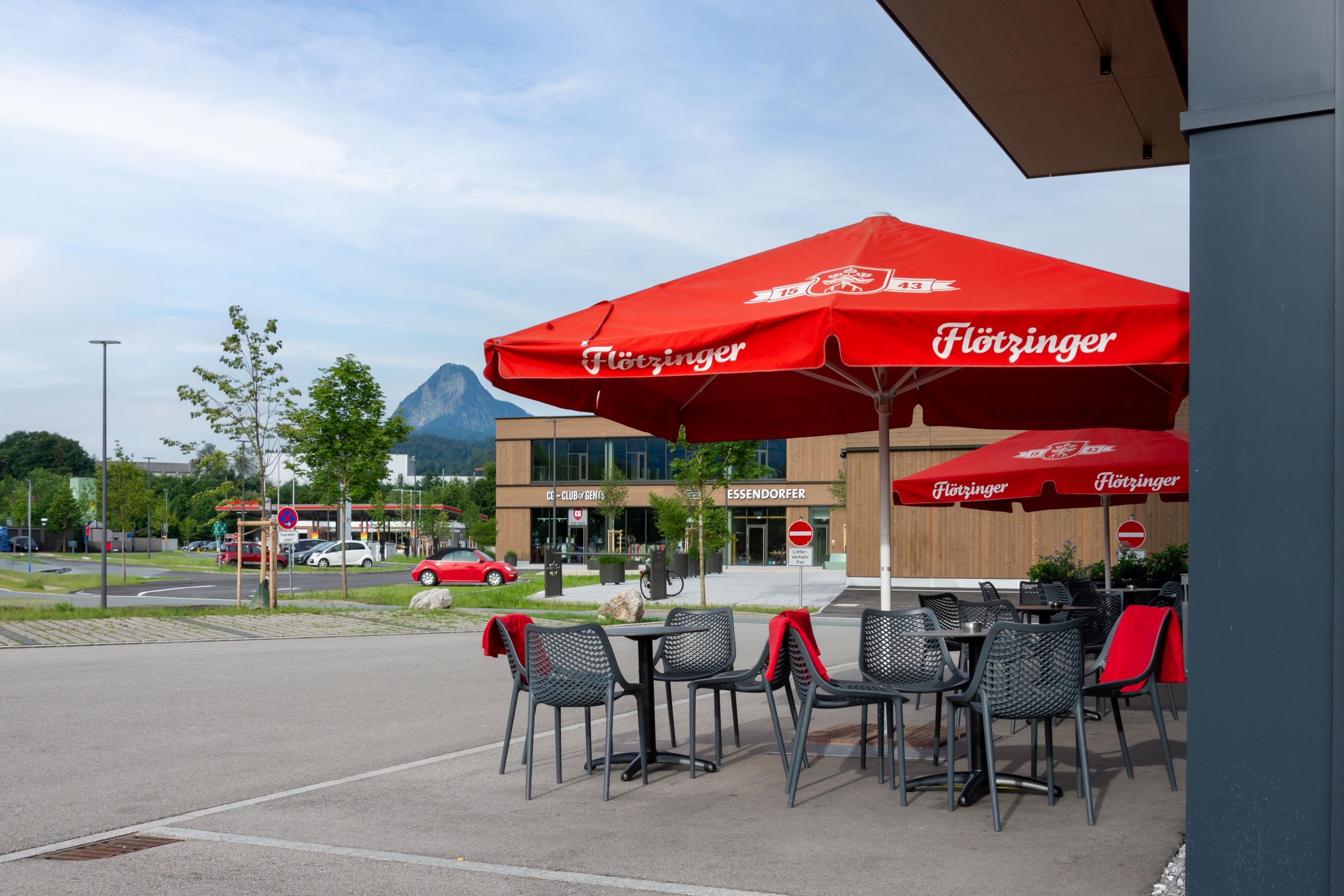 Sitzgelegenheiten im Freien mit roten Sonnenschirmen, ein modernes Gebäude mit Beschilderung und ein Berg im Hintergrund.