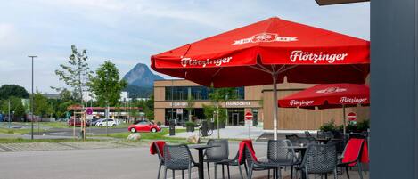 Sitzgelegenheiten im Freien mit roten Sonnenschirmen, ein modernes Gebäude mit Beschilderung und ein Berg im Hintergrund.