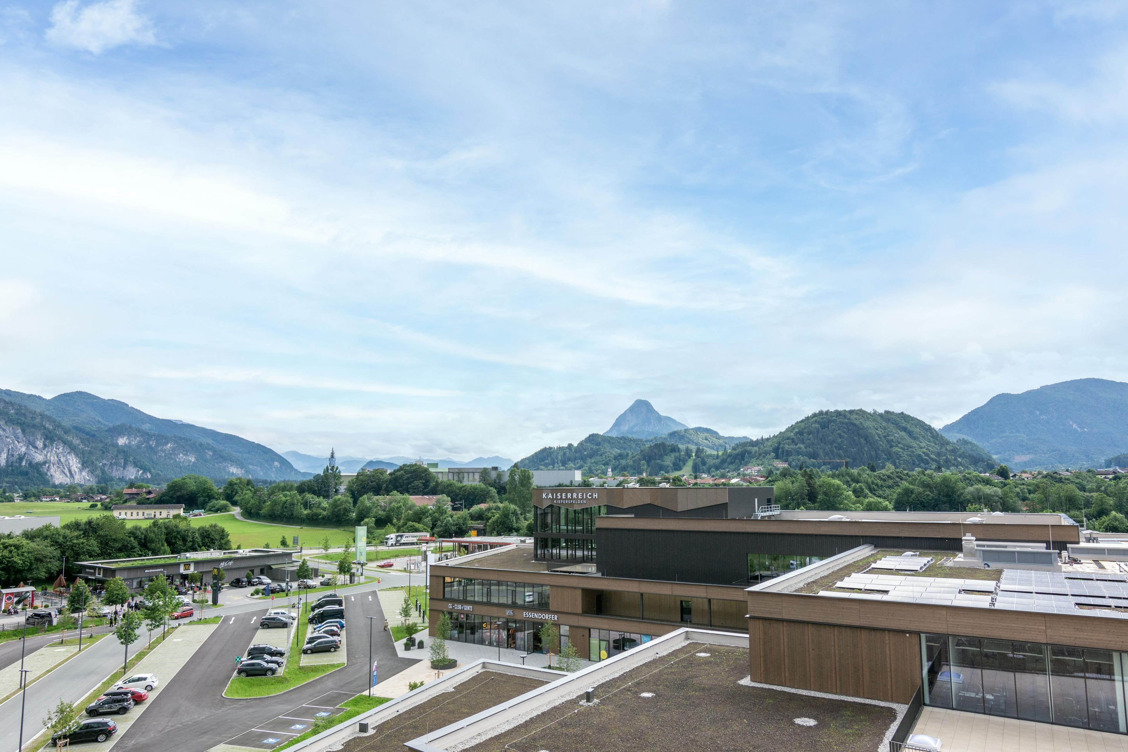 Edificio moderno con techo plano y paneles solares, rodeado de estacionamiento y vegetación, con montañas al fondo.