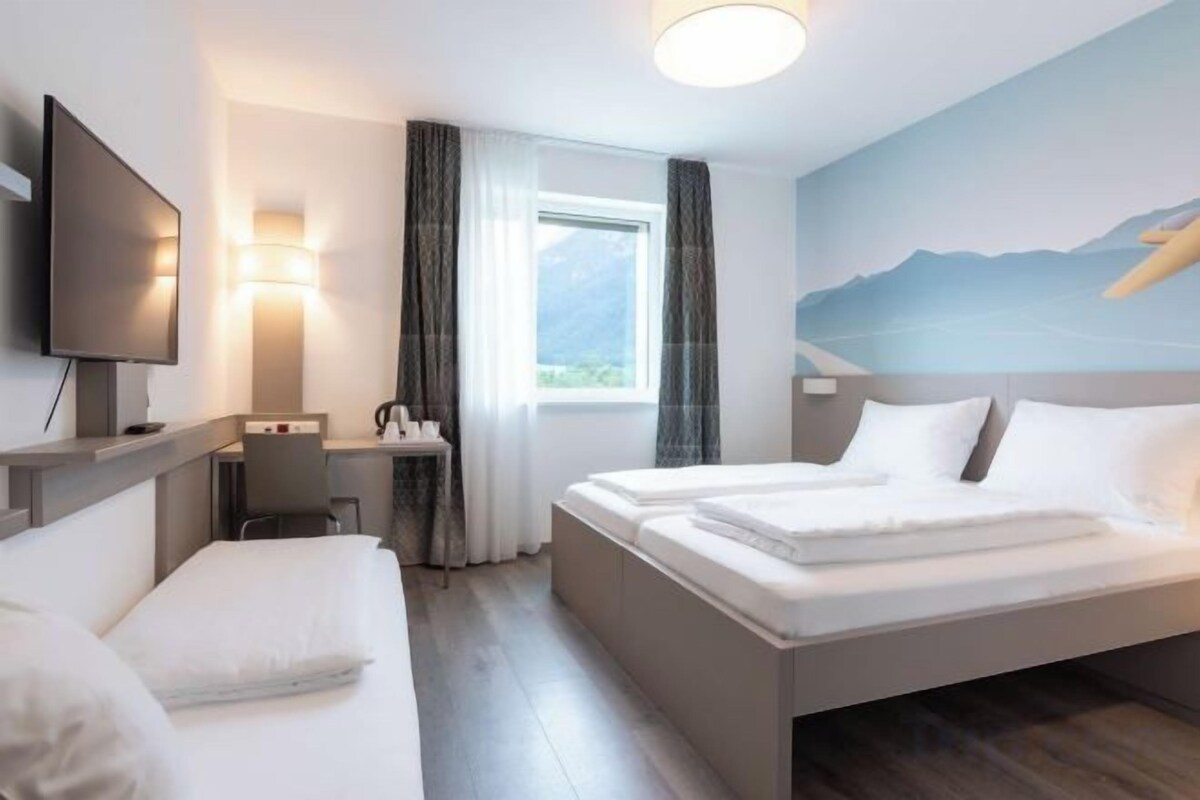 Una habitación de hotel con cama, escritorio, silla, televisor y ventana con cortinas.