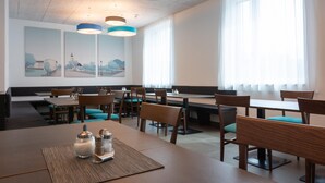 A modern dining area with multiple tables and chairs, large windows with sheer curtains, and artwork on the walls.