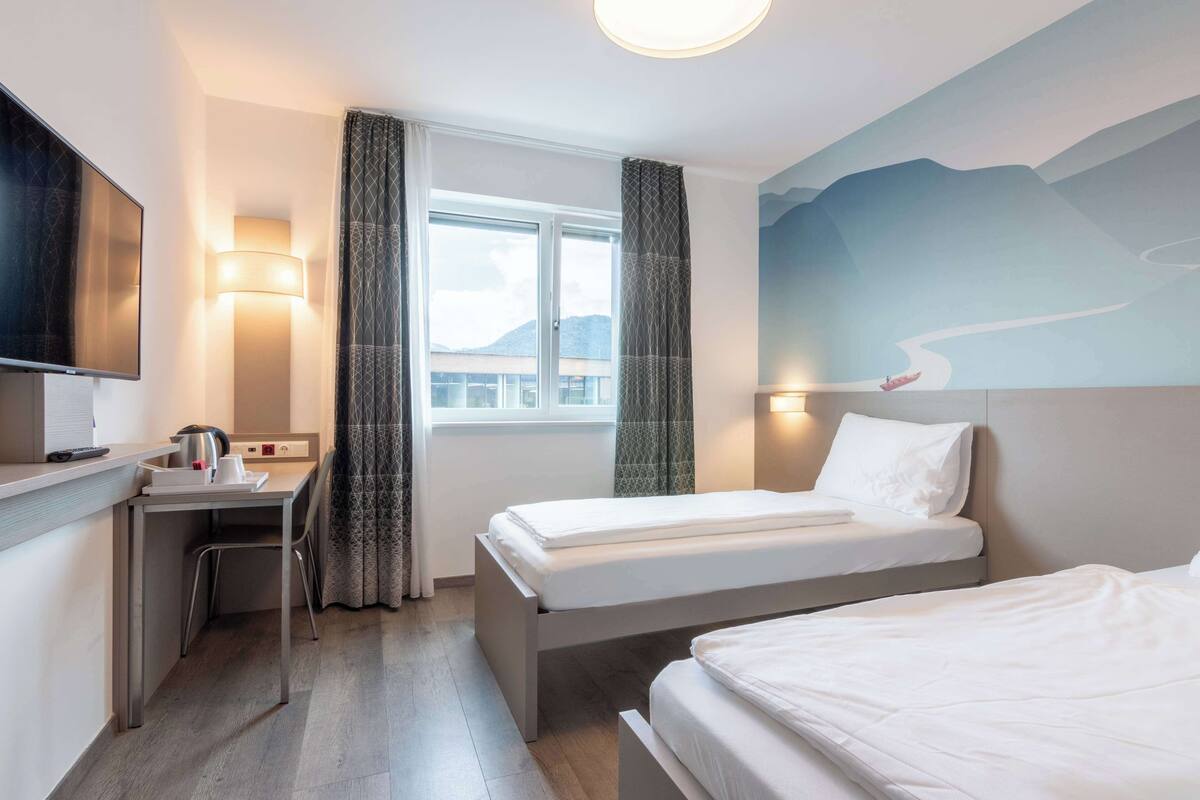 A hotel room with two beds, a desk, a TV, and a window with curtains.