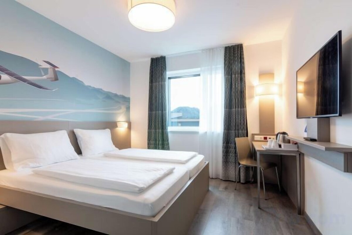Habitación de hotel con cama, escritorio, silla, televisor y ventana con cortinas.