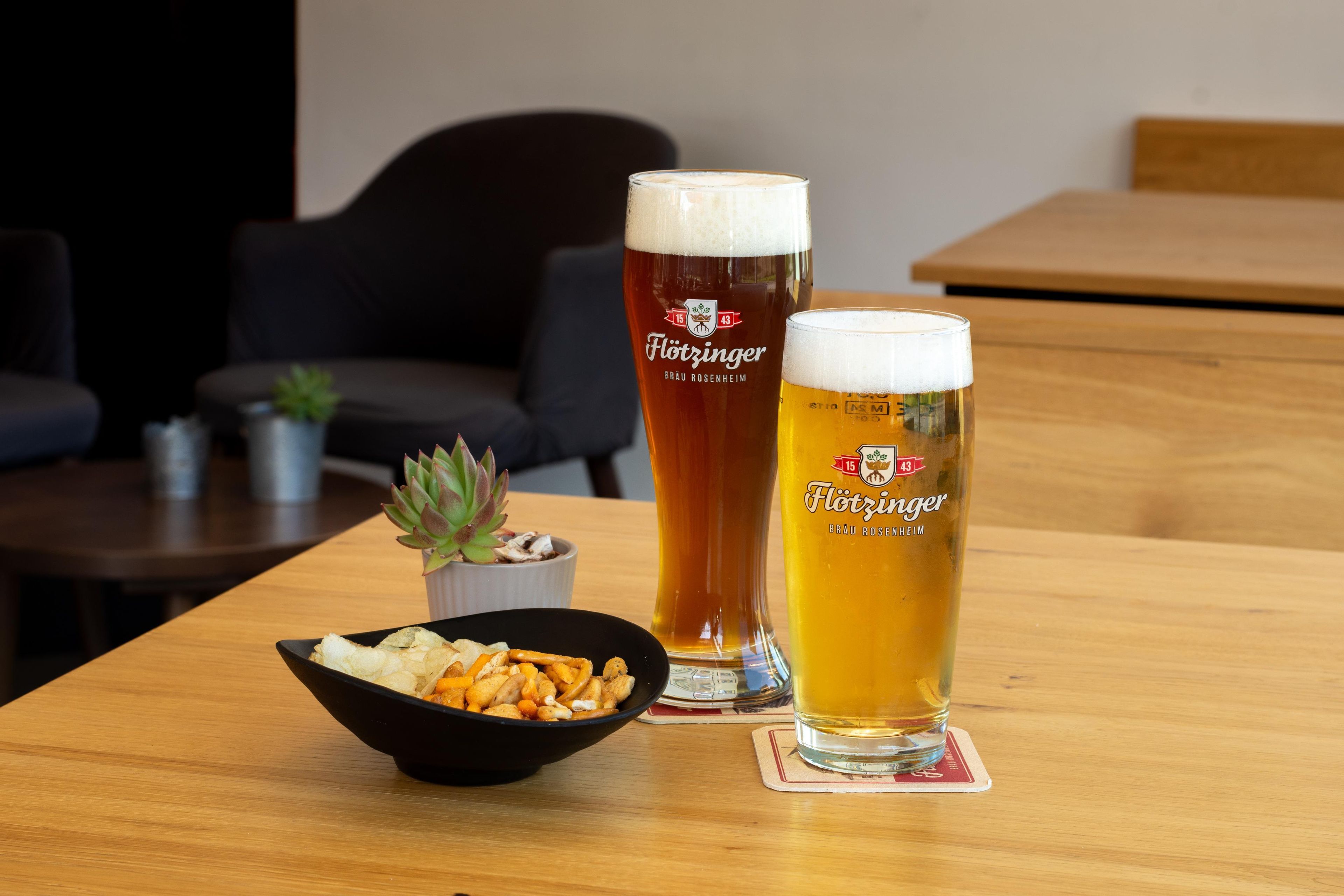 Dos vasos de cerveza sobre una mesa de madera con un cuenco de aperitivos y una pequeña planta en maceta.