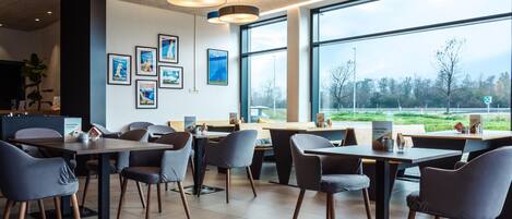 A modern dining area with tables and chairs, framed pictures on the wall, and large windows offering a view of the outdoors.