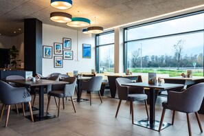 A modern dining area with tables and chairs, framed pictures on the wall, and large windows offering a view of the outdoors.