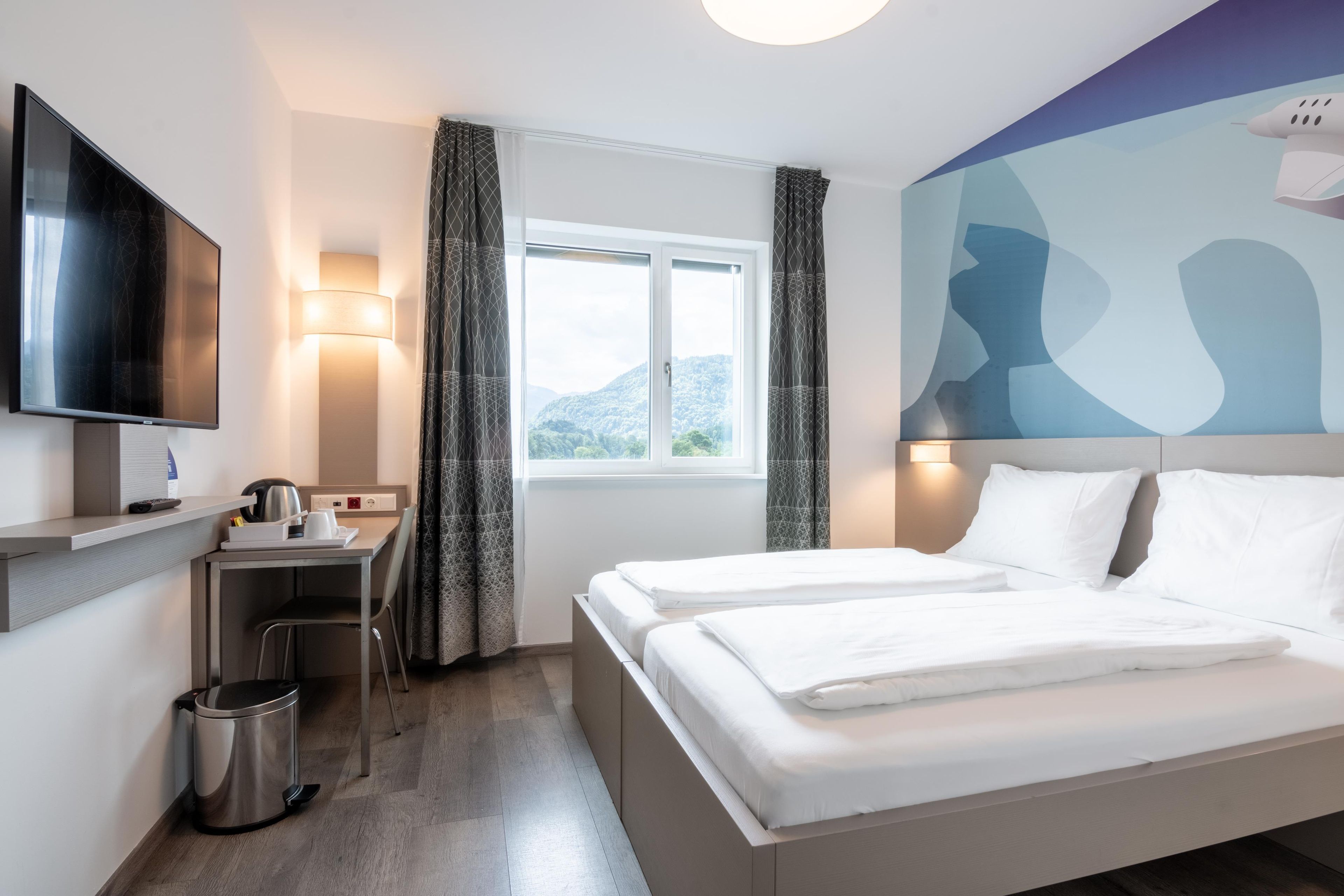 A hotel room with a bed, a desk with a kettle, a trash bin, a TV, and a window with curtains.