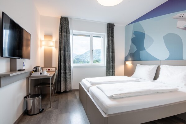 A hotel room with a bed, a desk with a kettle, a trash bin, a TV, and a window with curtains.