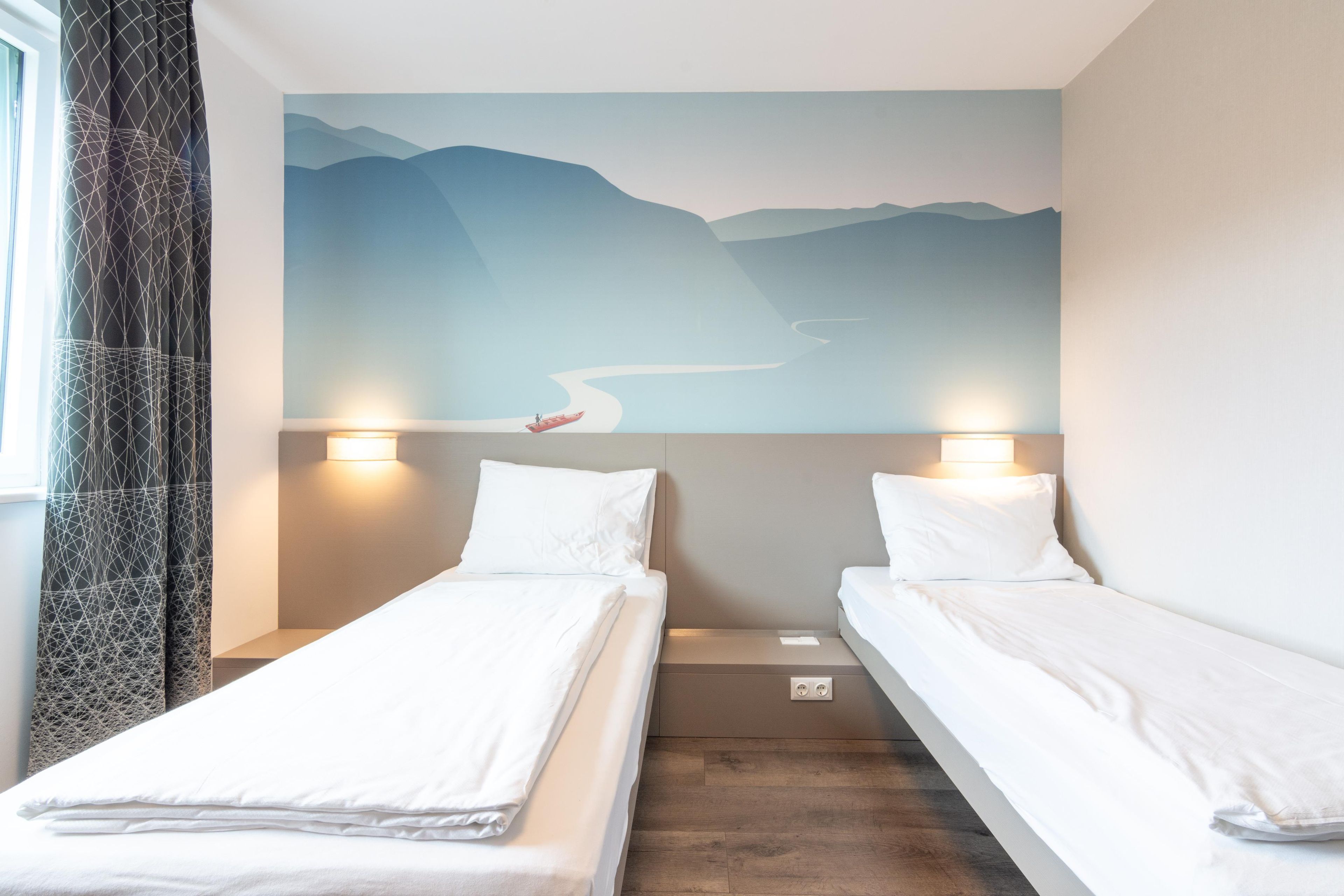 A hotel room with two single beds, a mural of a landscape on the wall, and a window with curtains.