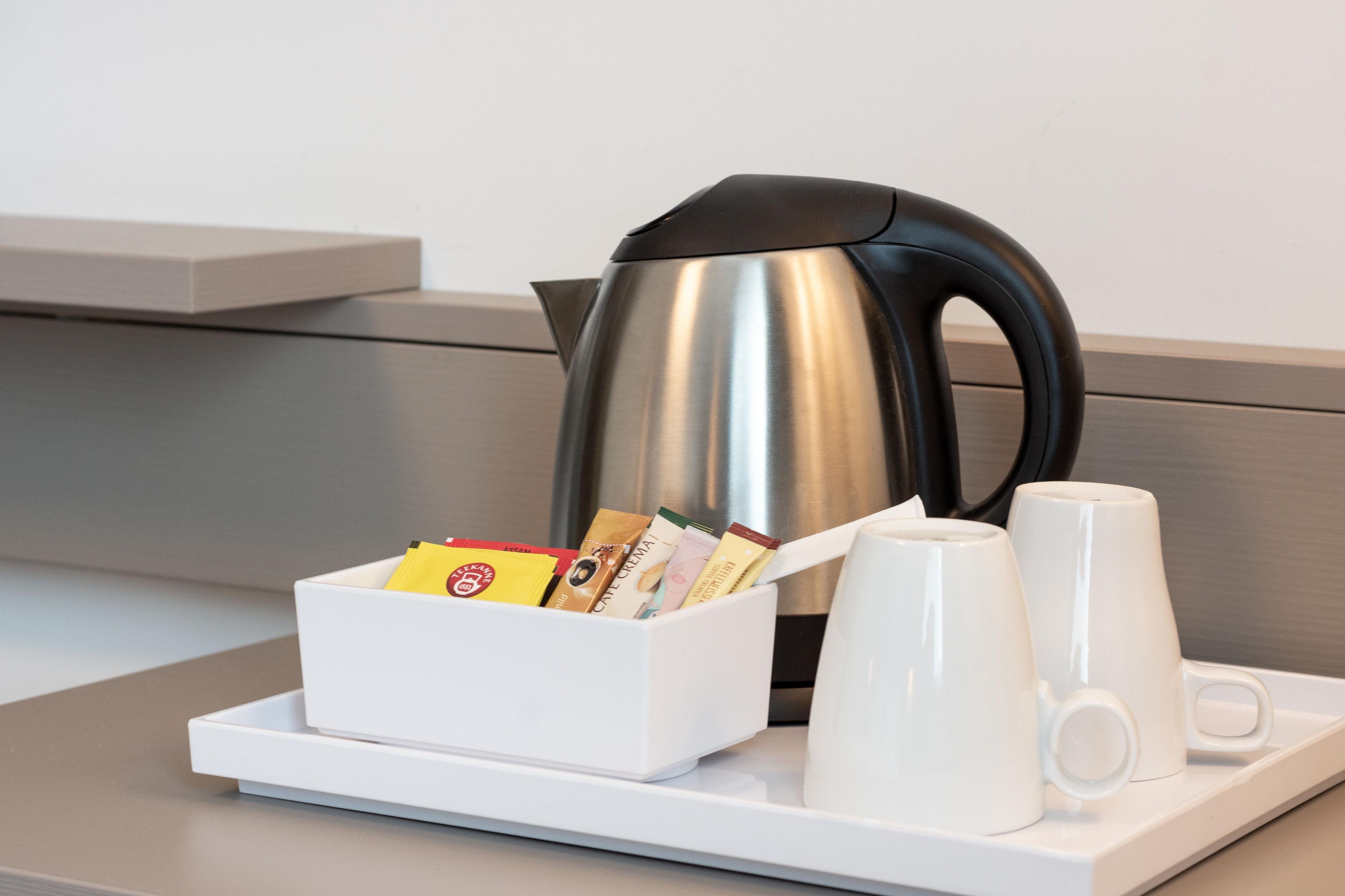 Una bandeja con una taza blanca, una taza de cerámica blanca, una caja blanca con cápsulas de café y una tetera de acero inoxidable.